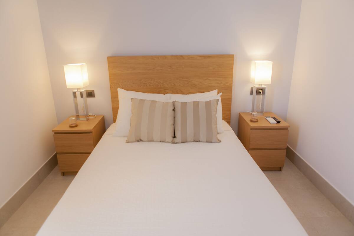 A neatly made bed with a wooden headboard, two bedside tables with lamps, and a plain wall.