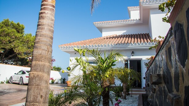 A two-story building with a tiled roof, surrounded by palm trees and a white wall.