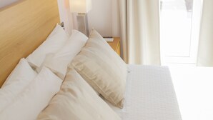 A neatly made bed with white linens and pillows, a bedside lamp, and a view of greenery through a window with curtains.