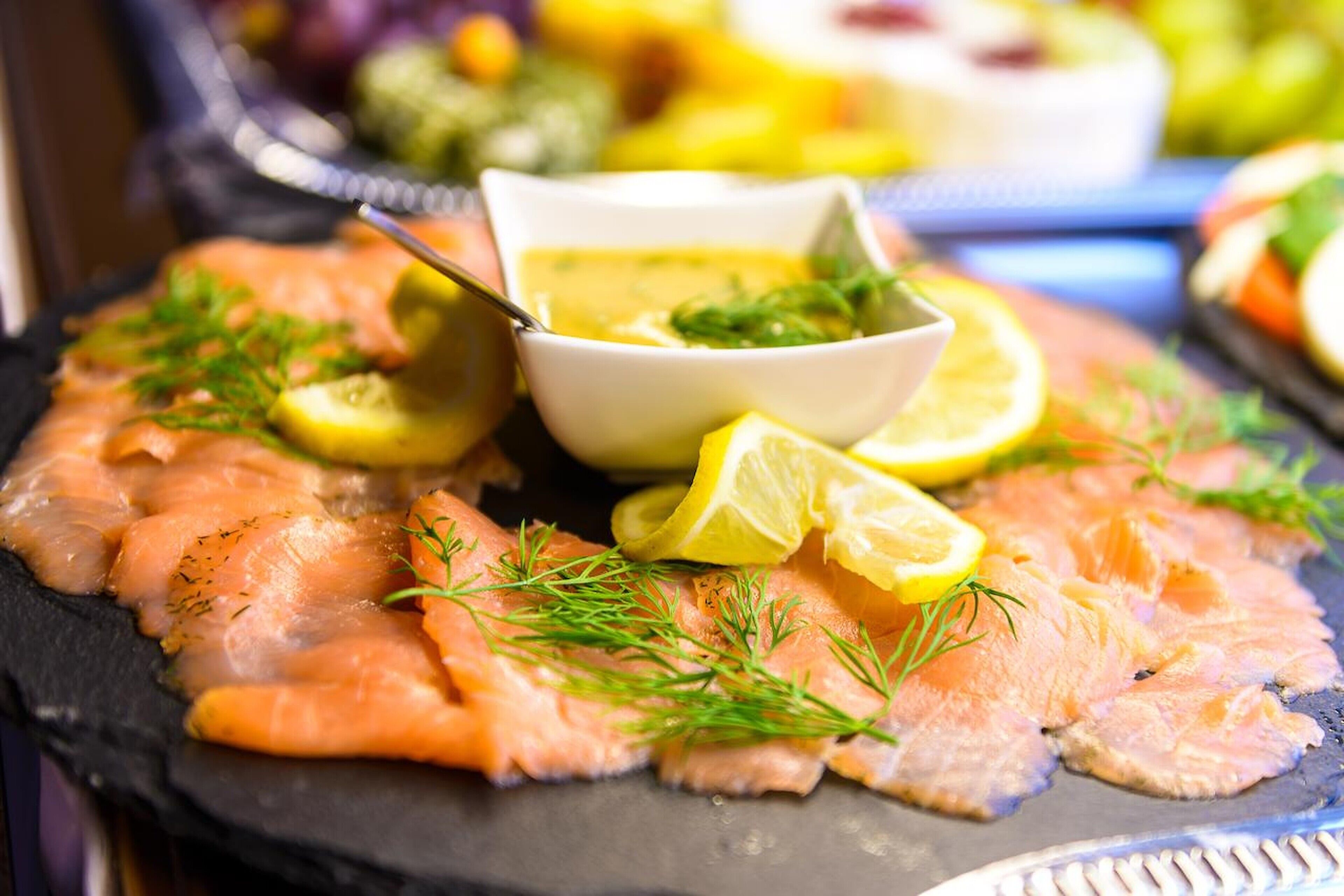 A platter of smoked salmon garnished with dill and lemon slices, accompanied by a small bowl of sauce.