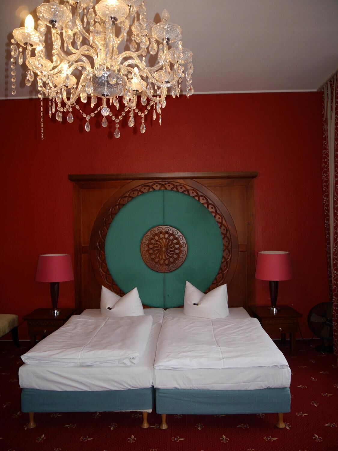 Een tweepersoonsbed met een blauwgroen hoofdeinde, twee nachtkastjes met lampen, en een kroonluchter.