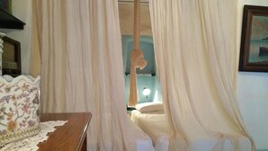A bedroom with a bed covered by sheer curtains, a bedside table with a floral-patterned vase, and a wall-mounted lamp. - Villa Via Savoia (Leros)