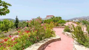 A paved pathway leading to a building with a red roof, surrounded by greenery and hills in the background. - Villa Via Savoia (Leros)