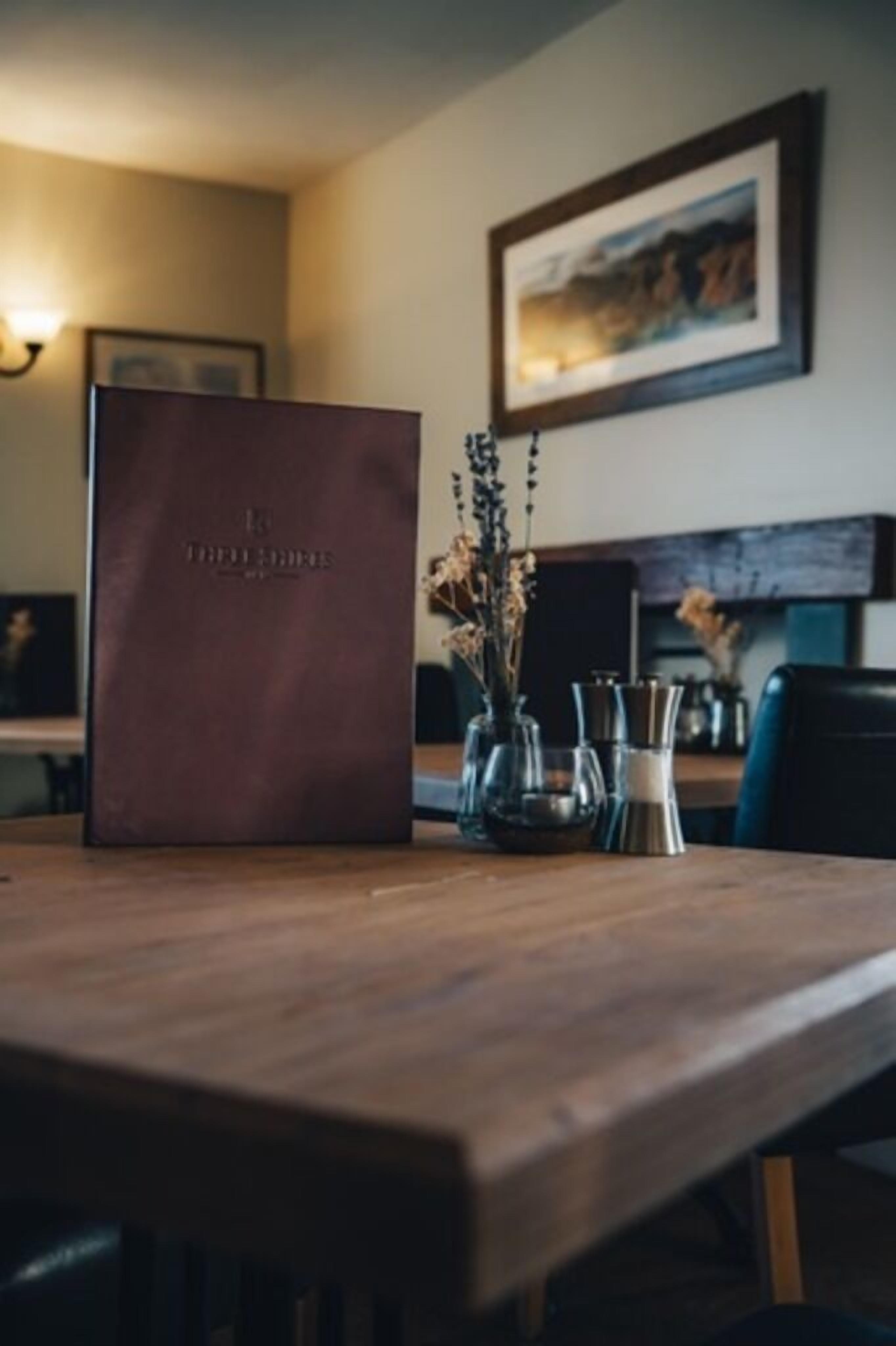 A wooden table with a brown leather-bound book, a vase with dried flowers, and a framed picture on the wall.