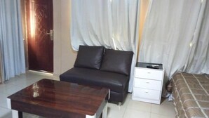 Una habitación de hotel compacta con una mesa de madera, un sofá con dos almohadas, una mesita de noche con cajones y una cama con colcha estampada.