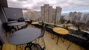 A rooftop terrace with multiple tables and chairs, offering a view of high-rise buildings and a distant mountain range. - hUB Antofagasta (Antofagasta)