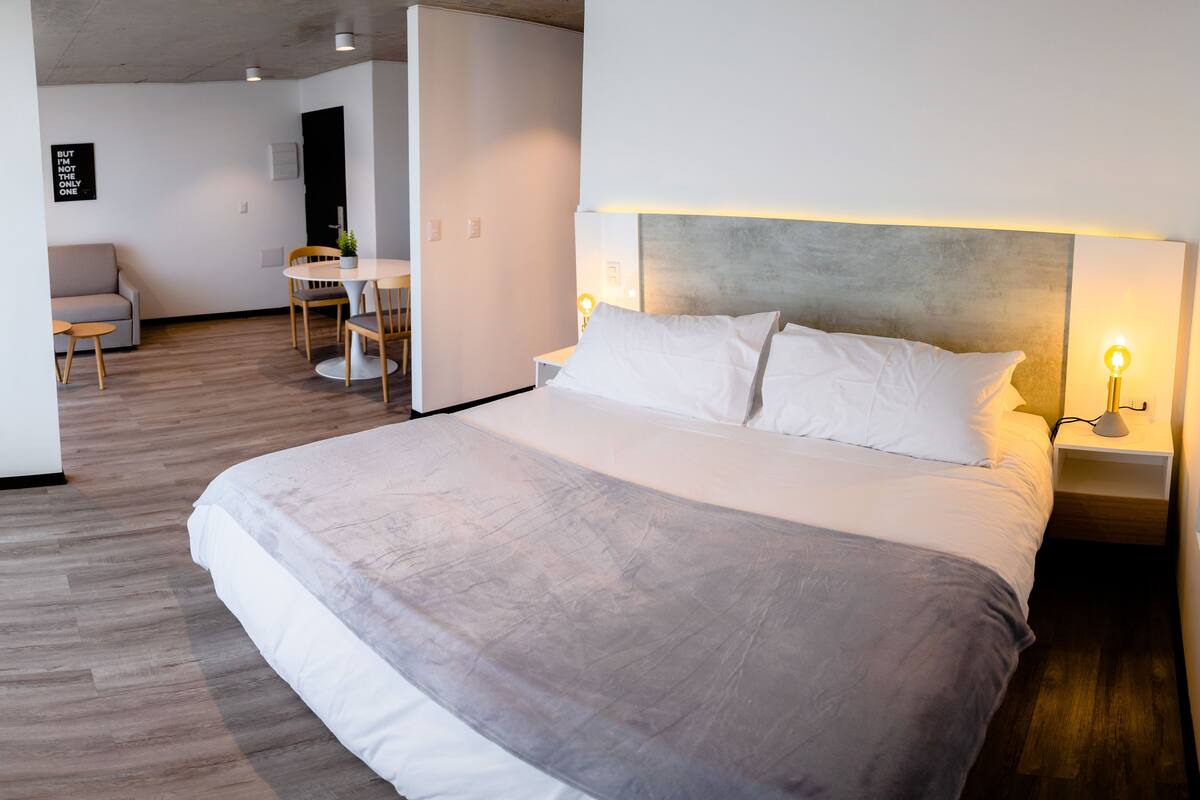 Una habitación de hotel moderna con una cama grande, mesitas de noche con lámparas, una pequeña mesa con sillas y vistas al área de estar contigua.