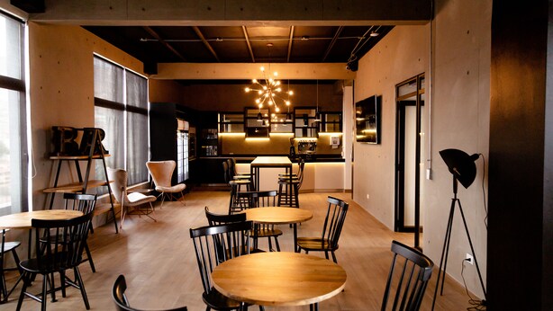 A modern interior with round wooden tables and black chairs, a kitchen area, and a large window providing natural light. - hUB Antofagasta (Antofagasta)