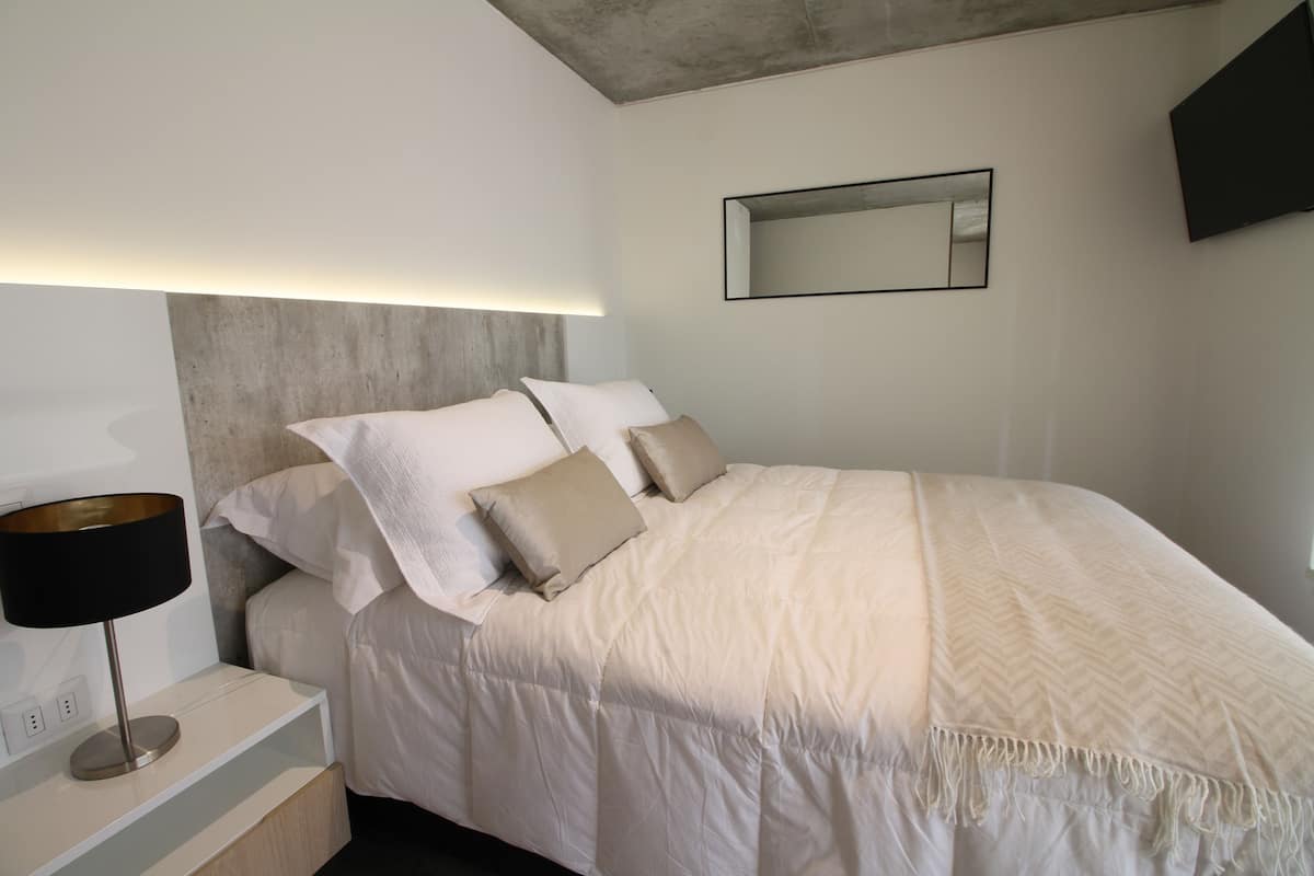 Un dormitorio moderno con cama, mesita de noche, lámpara y un espejo montado en la pared.