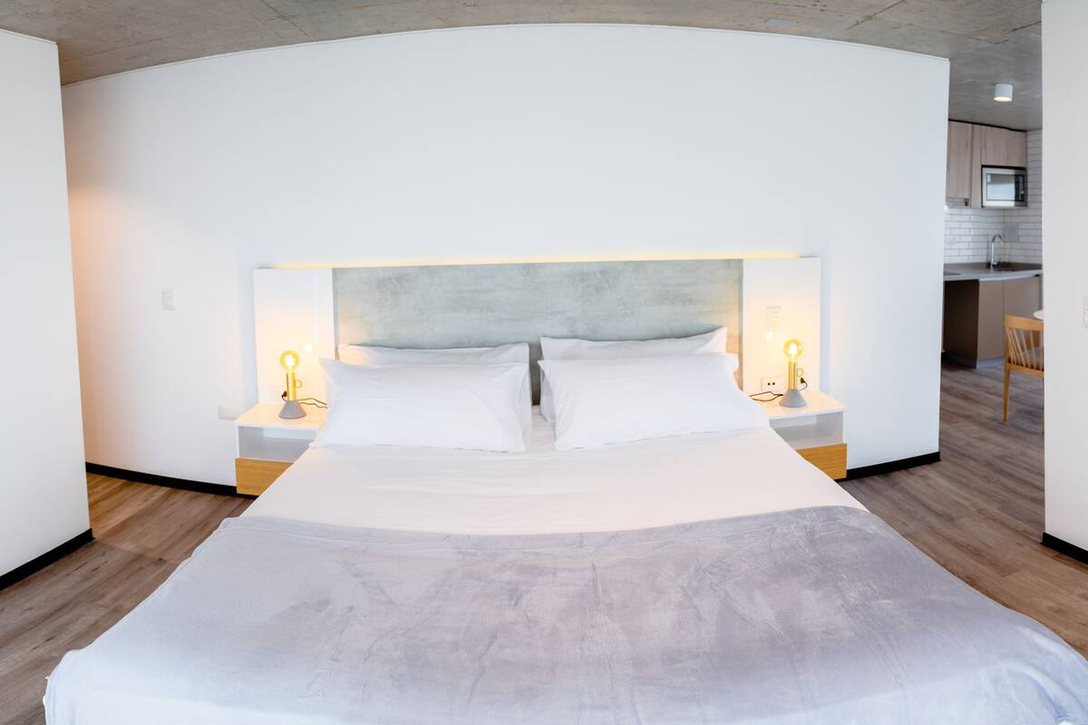 Una habitación de hotel moderna con distribución curva, una cama grande con ropa blanca, lámparas de noche y un televisor de pantalla plana montado en la pared.