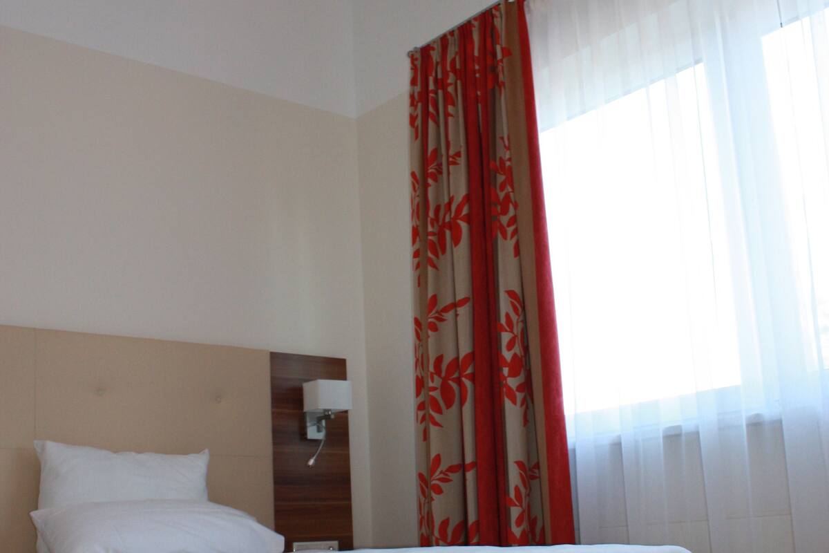 A bedroom with a wooden bed, white bedding, a nightstand with a lamp, and sheer curtains with a floral pattern.
