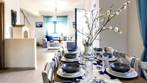 Ein Essbereich mit einem Tisch, der für sechs Personen gedeckt ist, Stühle, einer Vase mit Blumen und Blick ins Wohnzimmer.