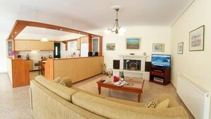 En moderne stue med sofa, sofabord og fjernsyn. Der er et køkkenområde med træskabe og et spisebord.