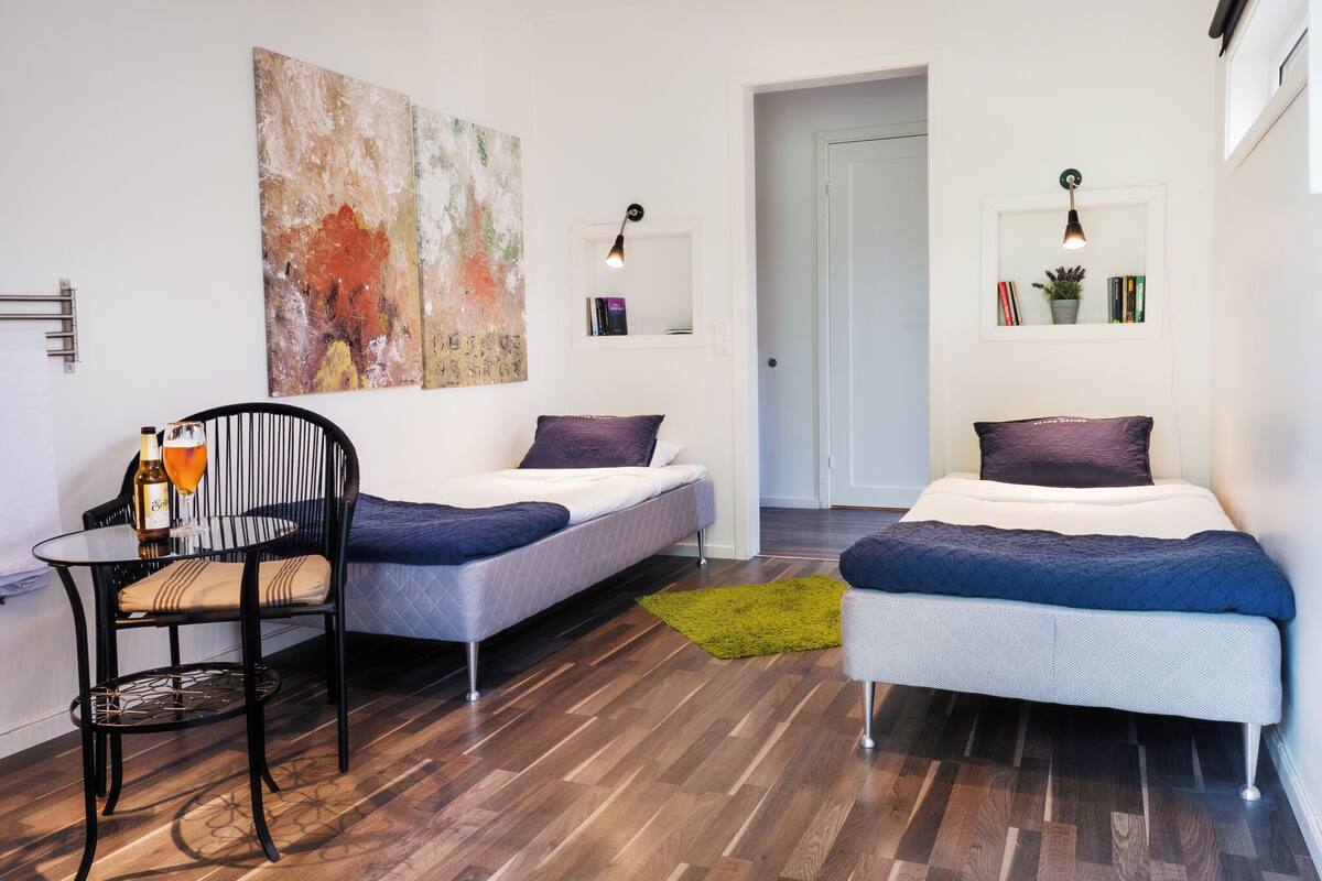 Ein modernes, kompaktes Zimmer mit zwei Einzelbetten, einem kleinen Tisch mit Stuhl, einem großen abstrakten Gemälde und einem Bücherregal mit Lampe.
