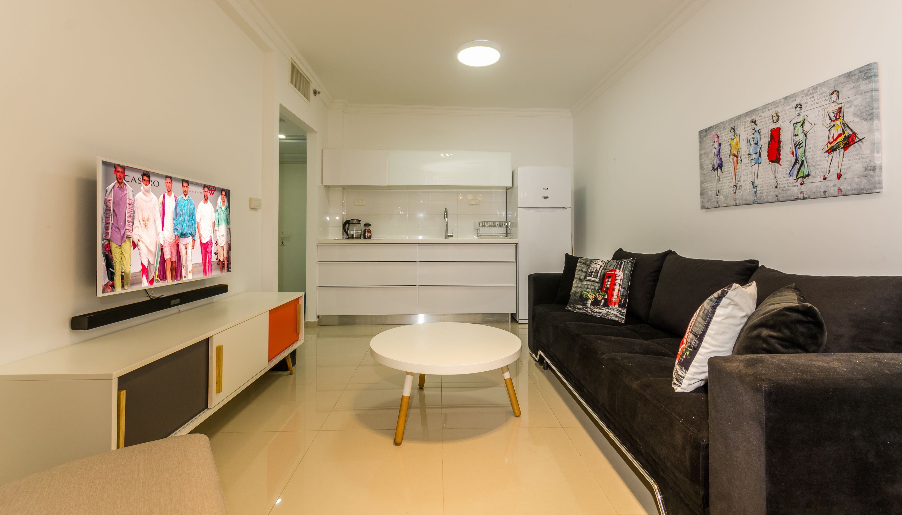 A modern living room with a flat-screen TV, a round coffee table, a black sofa, and a kitchen in the background.
