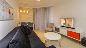 Uma sala de estar moderna com um sofá preto, uma mesa redonda branca, uma poltrona bege e uma TV sobre um móvel branco com detalhes laranja.