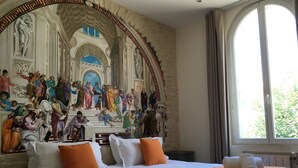 A hotel room with a large mural of a historical scene on the wall, a bed with white linens and orange pillows, and a window with curtains. - idealsejour (Cannes)
