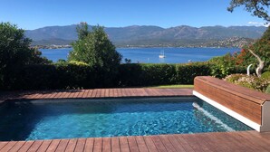 A swimming pool with a wooden deck, surrounded by greenery and a view of a lake with mountains in the background.