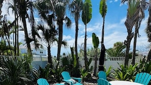 Une terrasse extérieure avec des chaises turquoise et une table ronde, entourée de palmiers et d’une clôture blanche.