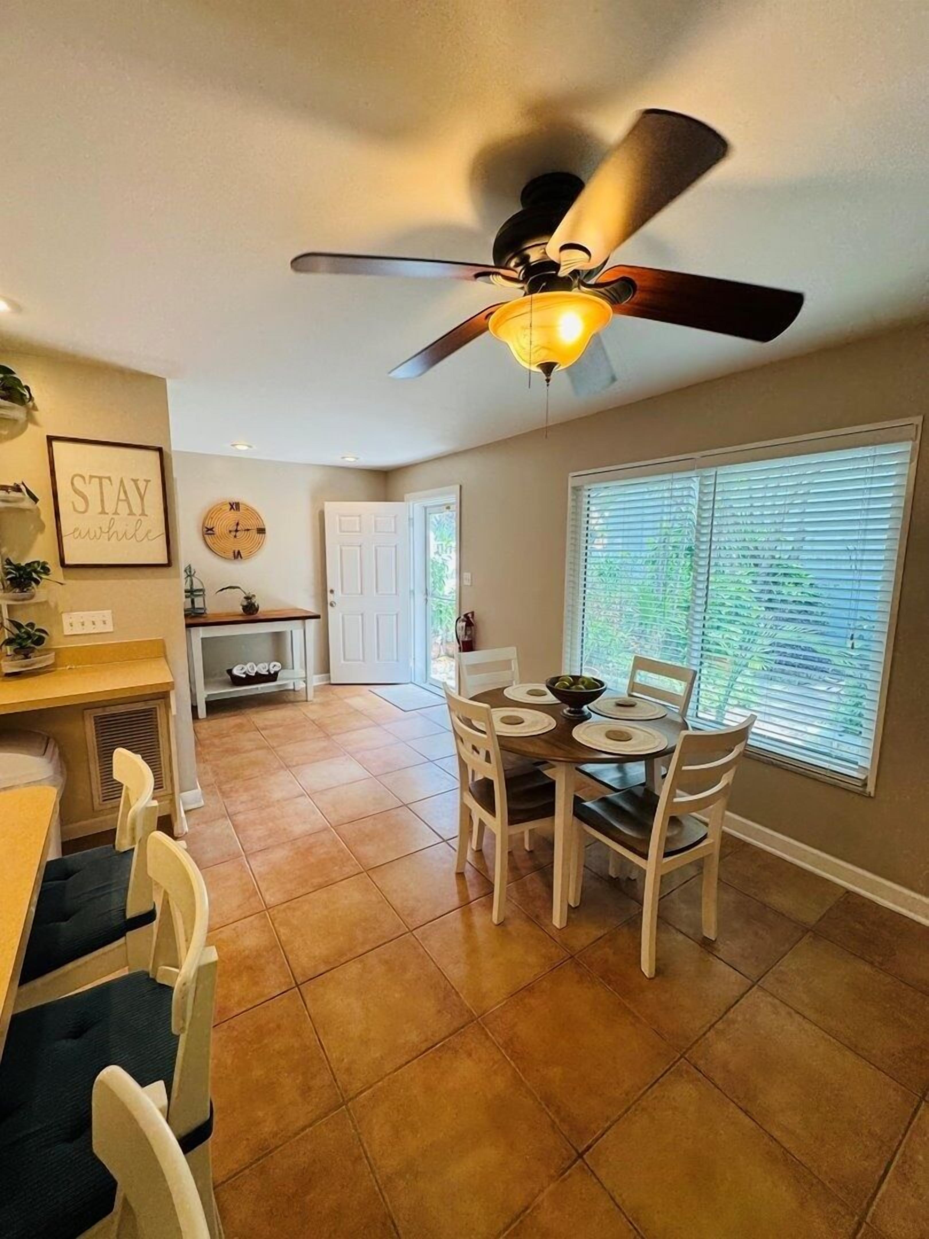 Un coin repas doté d’un ventilateur de plafond, d’une table à manger avec des chaises, d’un espace cuisine avec une porte et d’une fenêtre avec des stores.