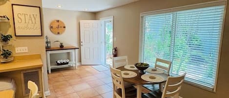 Un coin repas doté d’un ventilateur de plafond, d’une table à manger avec des chaises, d’un espace cuisine avec une porte et d’une fenêtre avec des stores.