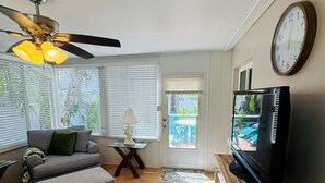 Un salon avec un ventilateur de plafond, un canapé, un pouf, un téléviseur posé sur un meuble et une horloge au mur.