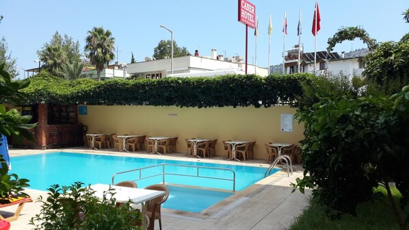 Una piscina con sillones de descanso y mesas, rodeada de follaje y un edificio con un cartel que dice 'CANER HOTEL'.