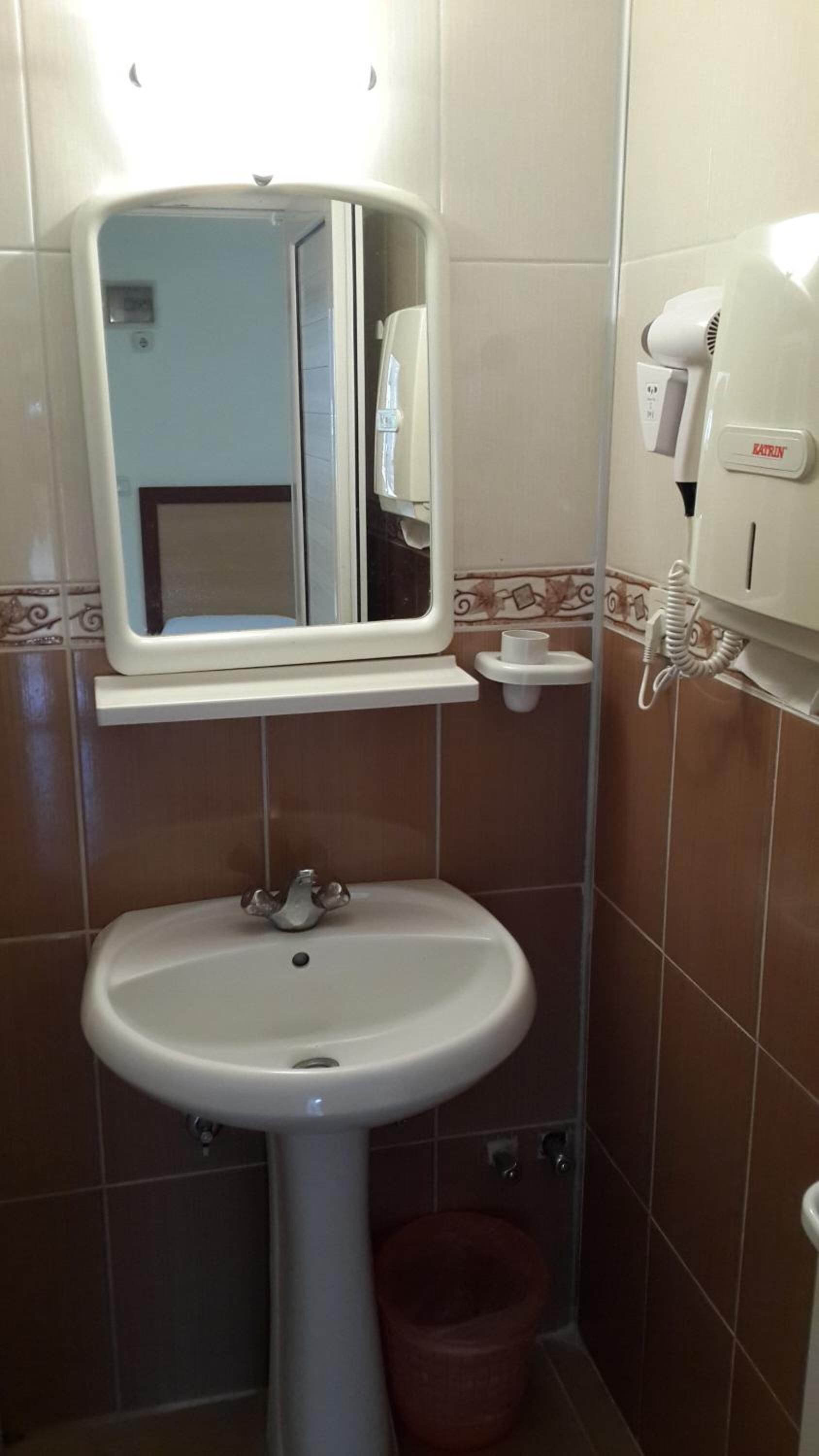 Un baño con lavamanos pedestal, espejo, secamanos y cubo de basura.