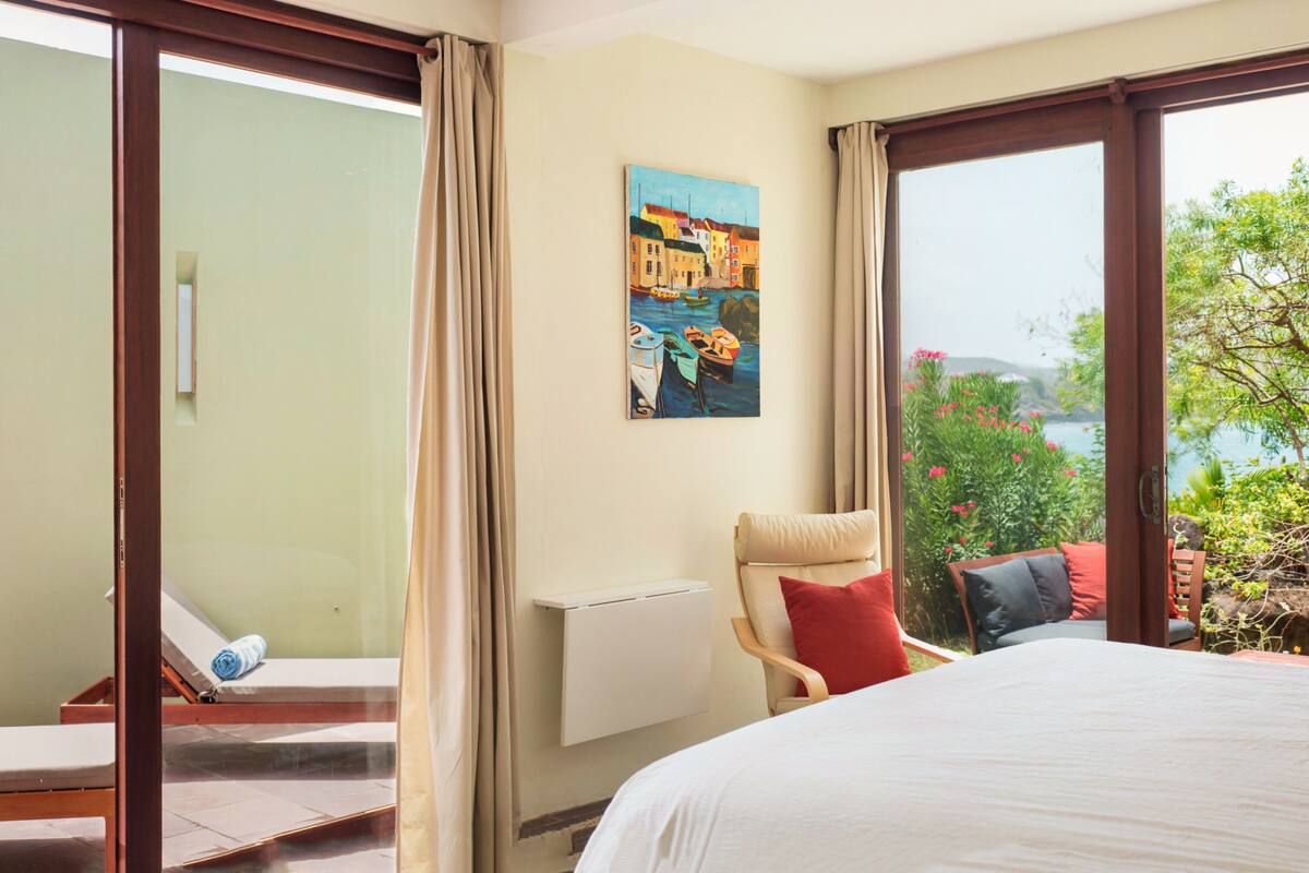 A hotel room with a bed, a chair, a painting, and a view of the sea.