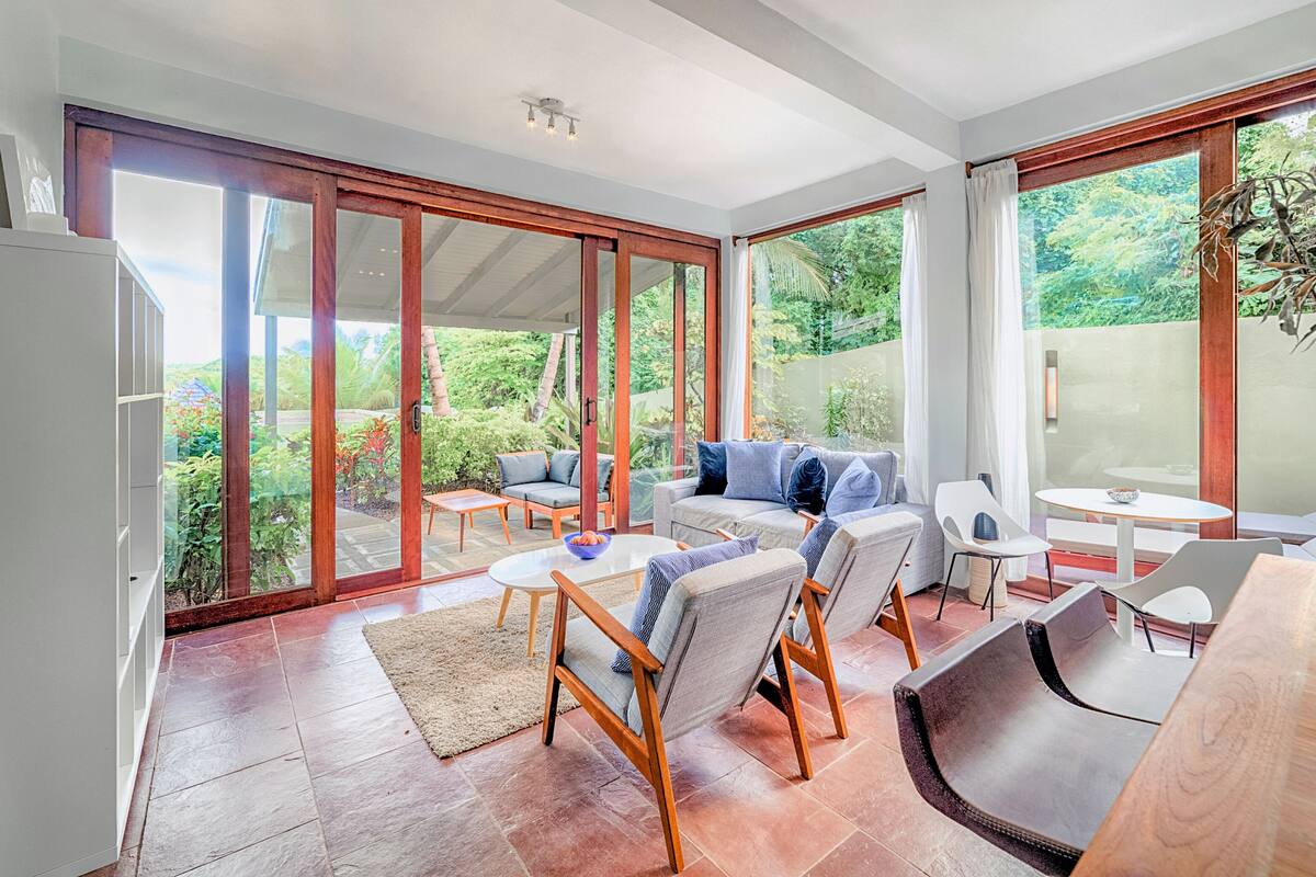 A spacious room with a large sliding glass door leading to a garden, a sofa set, a dining table, and a refrigerator.
