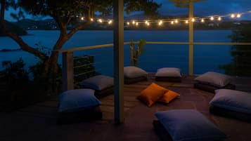 A terrace with outdoor seating and string lights, overlooking a body of water and hills.