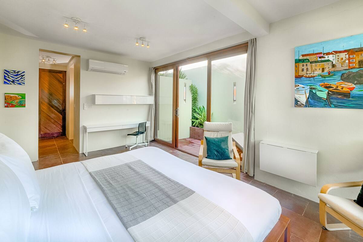 A modern hotel room with a large bed, a chair, a desk, and a painting of a coastal scene.