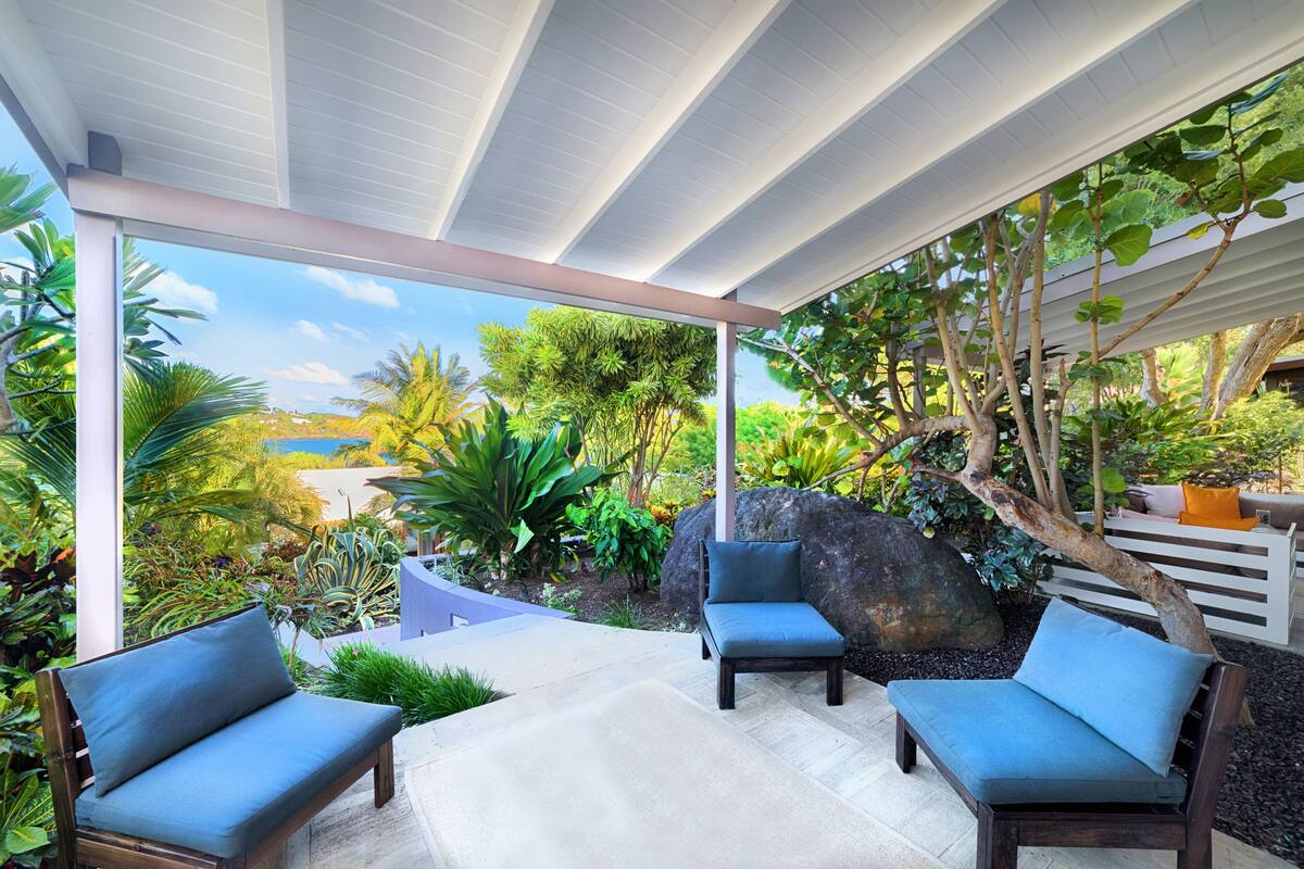 A covered outdoor seating area with two cushioned chairs and a bench, surrounded by lush greenery and a view of the landscape.