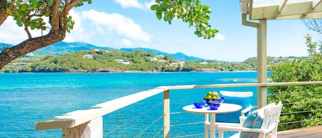 Una terraza de madera con una mesa y sillas que da a un mar azul cristalino y un paisaje montañoso.