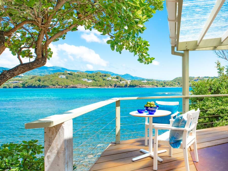 A wooden deck with a table and chairs overlooking a clear blue sea and a mountainous landscape.