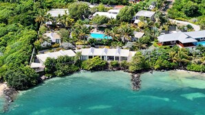 Oversigtsbillede af en kystresort med flere huse i bungalowstil, en swimmingpool og frodig vegetation.