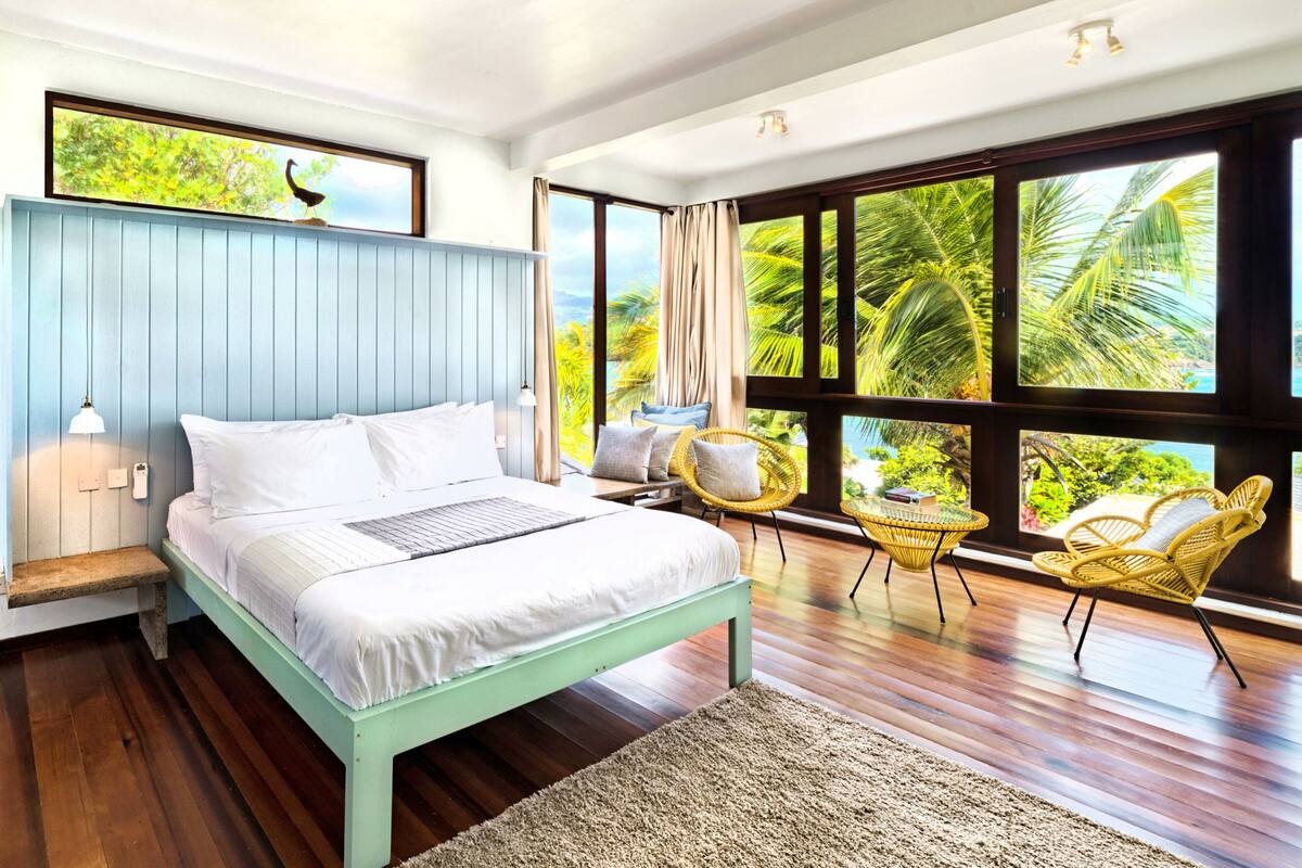 A modern bedroom with a bed, a nightstand, a lamp, a window with a view of palm trees, and a chair.