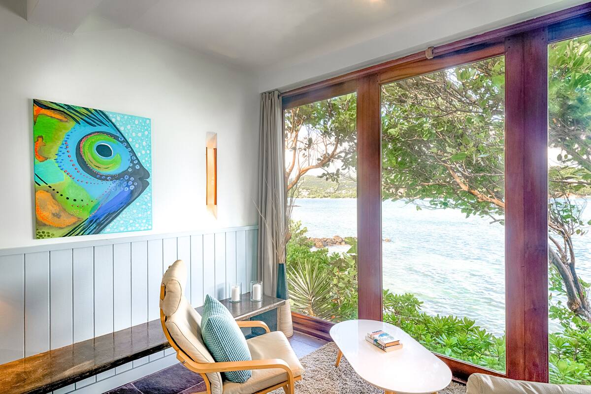 A room with a view of a body of water, a painting on the wall, and a seating area with a table.