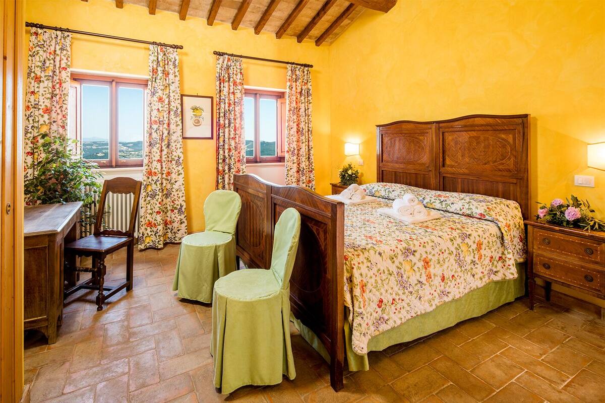 A bedroom with a wooden bed, two green chairs, a desk, and a view of the countryside through the window.