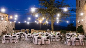 An outdoor dining area with stone flooring and tables set for a meal, surrounded by stone walls and a tree. - Castello di Titignano (Orvieto)
