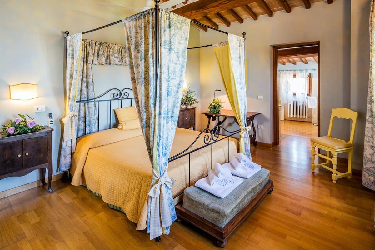 A four-poster canopy bed with a wooden bench, a bedside table with a lamp, and a chair in a room with wooden flooring and a view of a balcony.