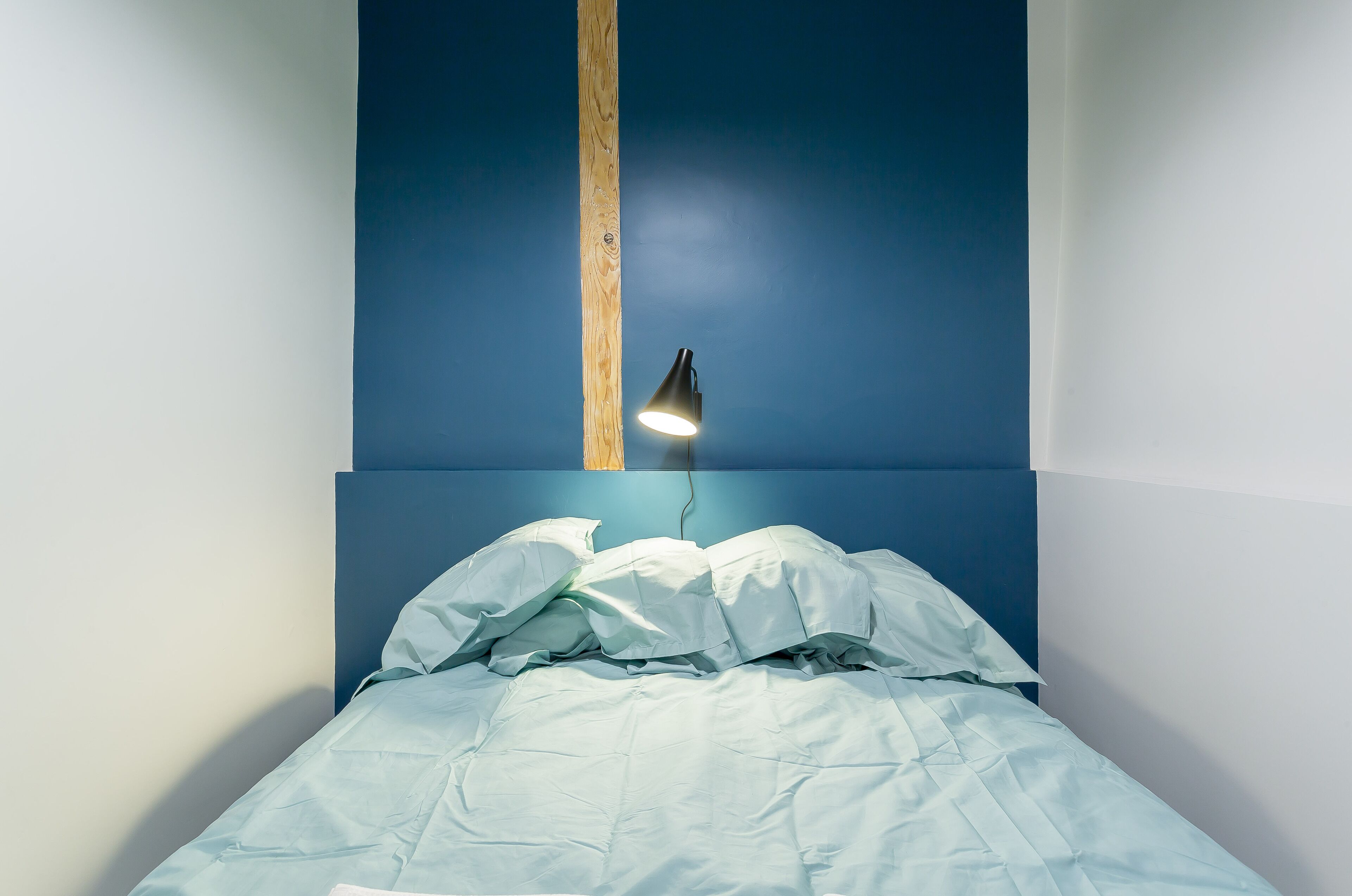 Un lit avec une literie blanche et une tête de lit en bois, adossé à un mur bleu. Un seul luminaire est suspendu au plafond.