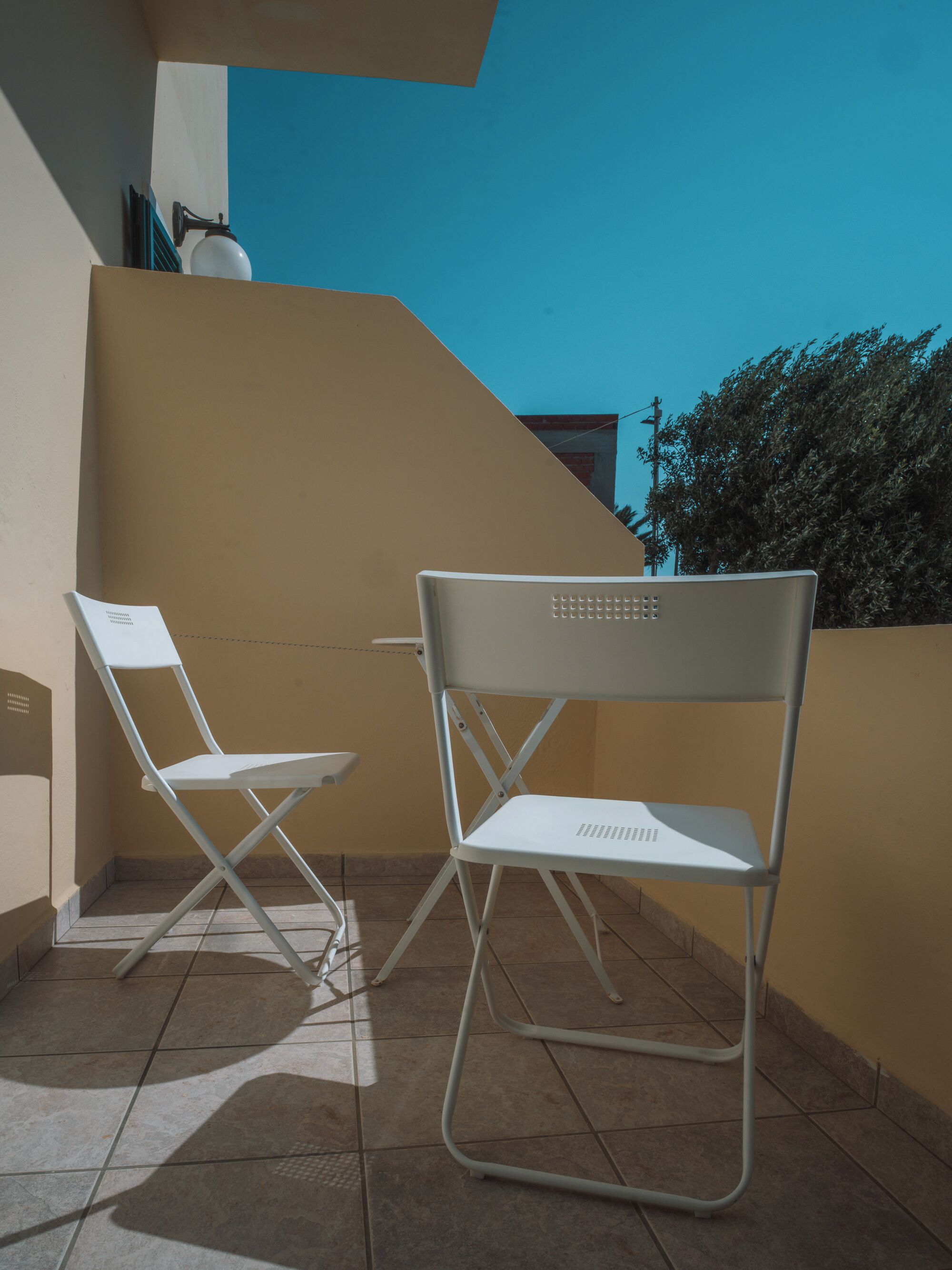 Two white folding chairs on a tiled balcony with a beige wall and a clear blue sky in the background.