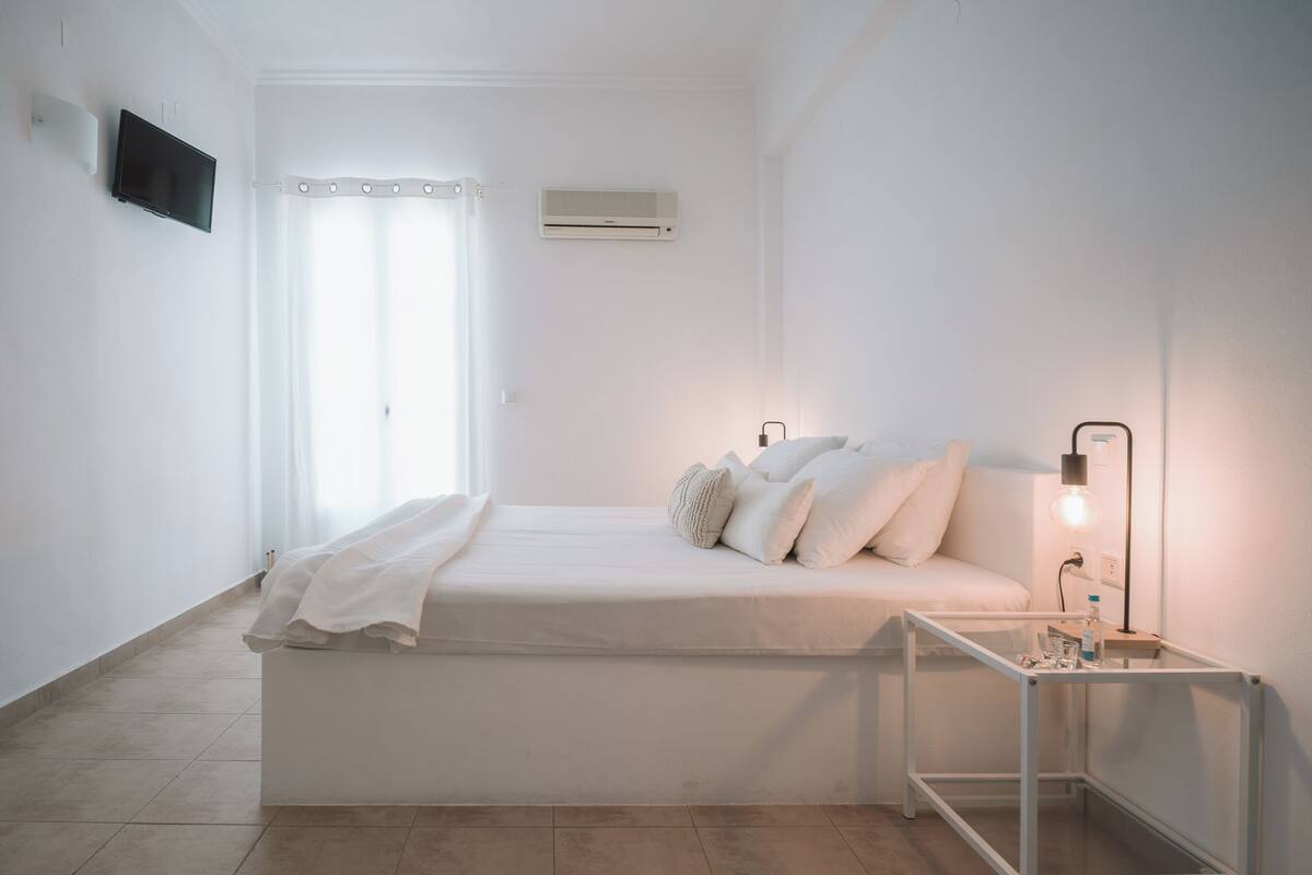 A minimalist bedroom with a bed, a bedside table with a lamp, a TV, and an air conditioning unit.