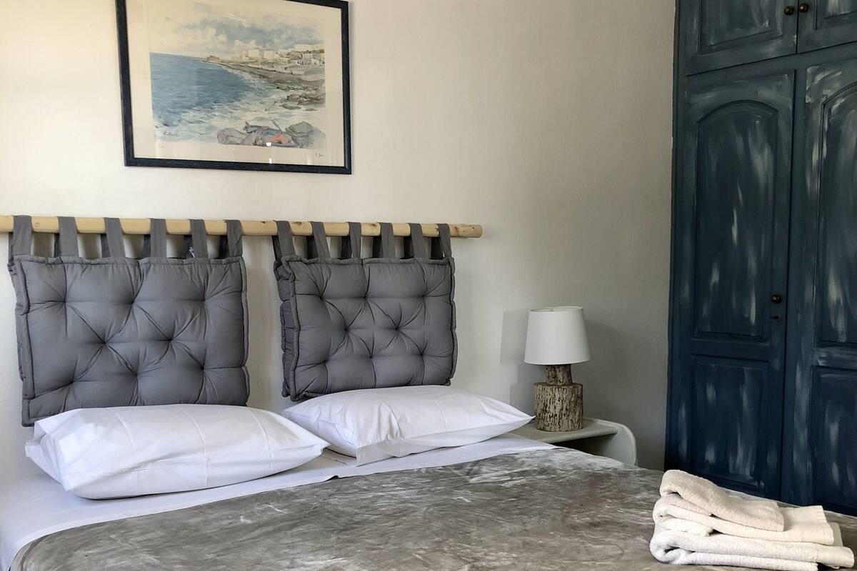 A neatly made bed with a grey duvet, white pillows, and a folded towel on the side. A framed picture hangs above the bed, and a lamp is on a bedside table.
