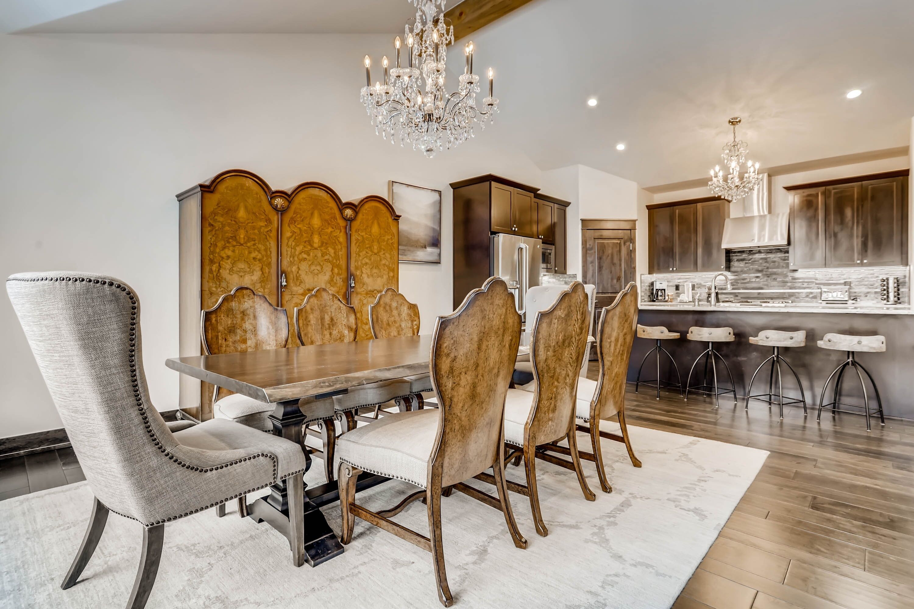 Un comedor con mesa de madera y sillas tapizadas, una lámpara de araña y una cocina con armarios oscuros y encimera de mármol.