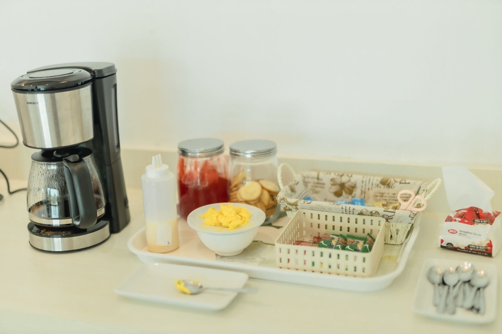 Un taulell de cuina amb una cafetera, pots d'ingredients, un bol d'ous, un plat de tapes i una caixa de bosses de te.