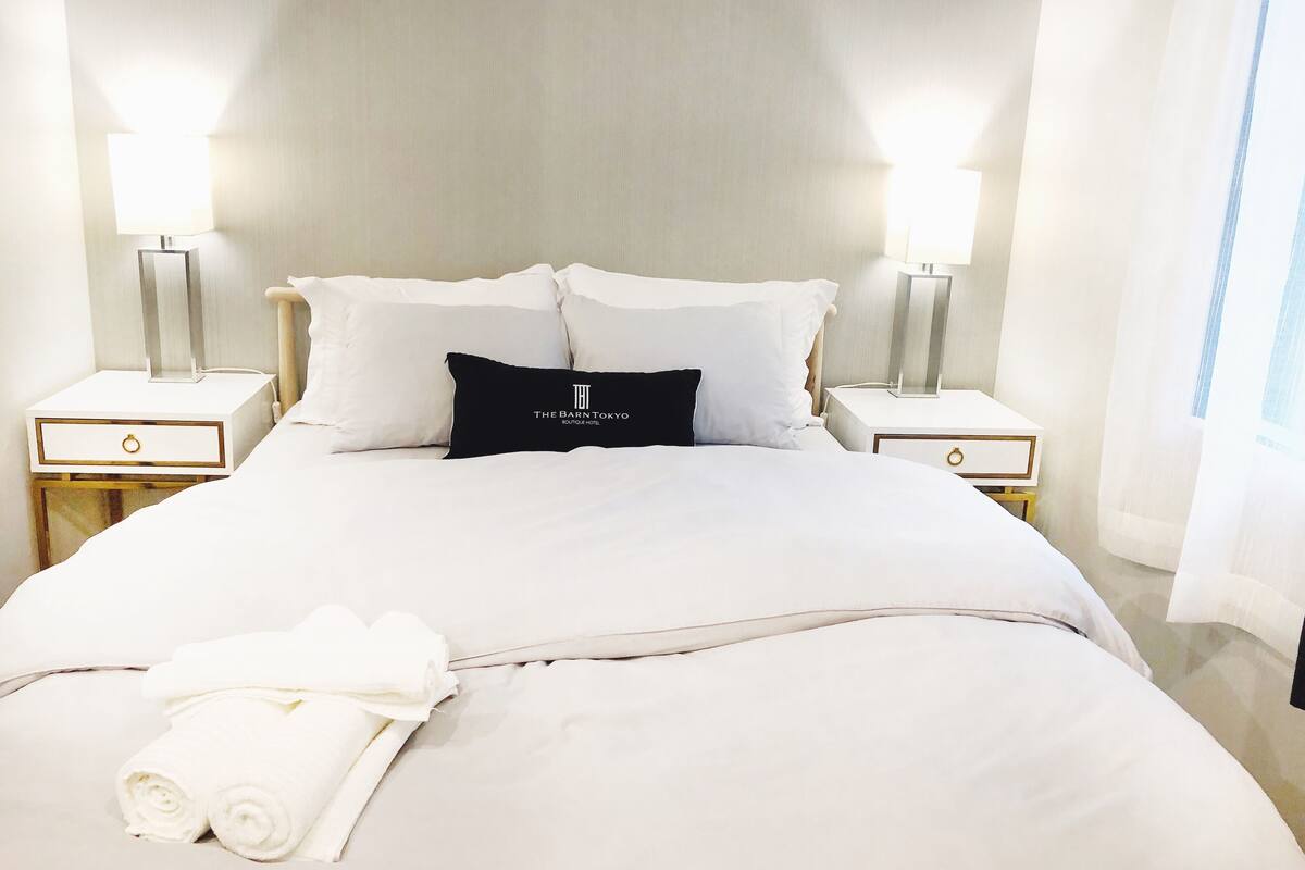 A neatly made bed with white linens and a black pillow with text, two bedside tables with lamps, and a window with sheer curtains.