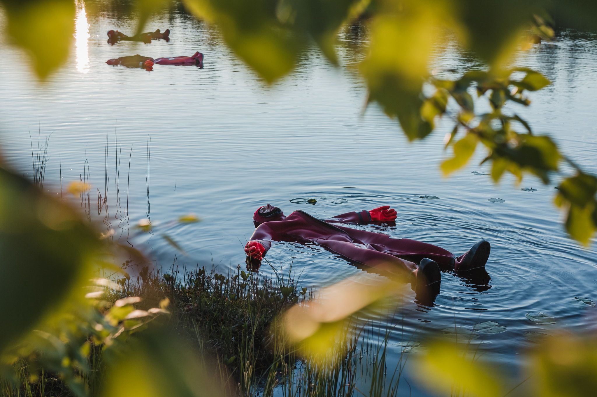 Une personne vêtue d’une combinaison de plongée rouge flotte sur une étendue d’eau calme, et les arbres et l’herbe environnants se reflètent à la surface.