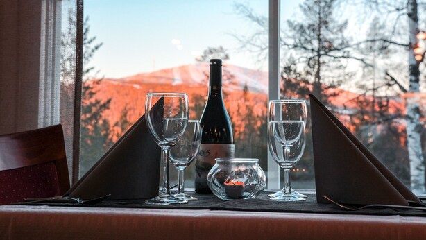 A table set with wine glasses, a bottle, and a candle, with a view of a mountain through a window. - Hotel Ylläsrinne (Kolari)